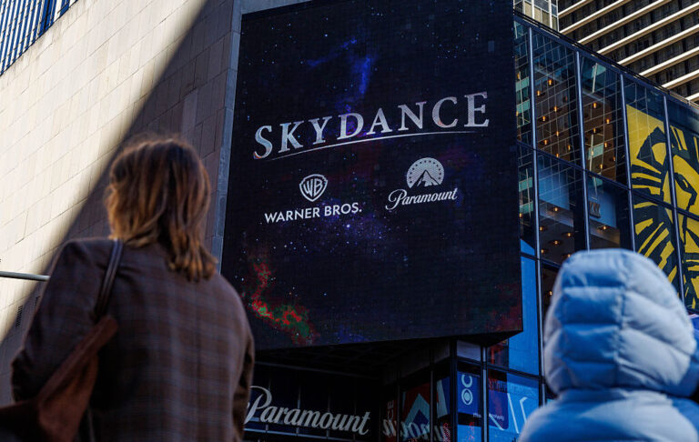 A black billboard that has the Skydance, WB, and Paramount logos. People walking past the billboard are visible in the foreground.