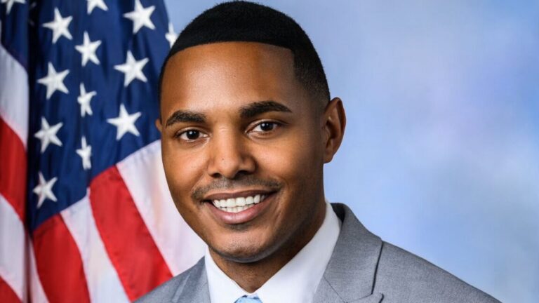 A man smiles in a suit in front of an American flag